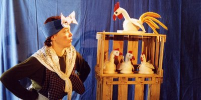 Eine Frau mit Schürze, Halstuch und Stirnband begutachtet einen Holzkäfig mit drei Stoffhühnern und einem Hahn.