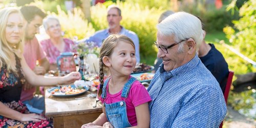 Eine Großfamilie feiert eine Gartenparty - im Vordergrund sitzt ein kleines Mädchen auf dem Schoß von ihrem Großvater