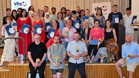 Regionalverbandsdirektorin Dr. Carolin Lehberger (2.v.r. sitzend) und vhs-Direktorin Nicole Boissier (links daneben), freuen sich gemeinsam mit 23 der Absolventinnen und Absolventen, ihren Lehrkräften und zuständigen vhs-Mitarbeitenden über den bestandenen Schulabschluss. Alle sitzen oder stehen in vier Reihen auf der Bühne im vhs-Zentrum.