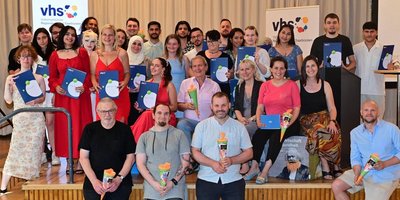 Regionalverbandsdirektorin Dr. Carolin Lehberger (2.v.r. sitzend) und vhs-Direktorin Nicole Boissier (links daneben), freuen sich gemeinsam mit 23 der Absolventinnen und Absolventen, ihren Lehrkräften und zuständigen vhs-Mitarbeitenden über den bestandenen Schulabschluss. Alle sitzen oder stehen in vier Reihen auf der Bühne im vhs-Zentrum.
