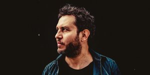 Vor schwarzem Hintergrund: Reto Burrell, ein dunkelhhariger Mann mit kurzem, dunkelbraunem Bart, blickt zur Seite.
