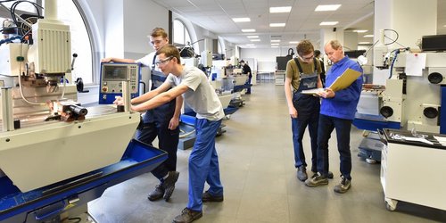 Ein Lehrer erklärt einem Schüler etwas anhand von Unterlagen. Weitere Schüler stehen an Maschinen. 