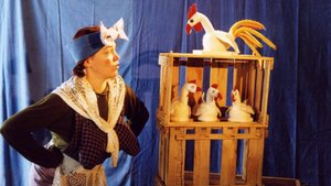Eine Frau mit Schürze, Halstuch und Stirnband begutachtet einen Holzkäfig mit drei Stoffhühnern und einem Hahn.