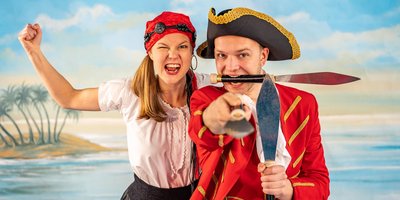 Jakob Mathias und seine Assistentin in Piratenkleidung vor dem Szenenbild einer Insel.