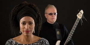 Vor schwarzem Hintergrund: Eine Frau mit dunklem, lockigem Haar blickt in die Kamera, hinter ihr ein älterer Mann mit Gitarre und getönter Brille.