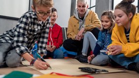 Boys'Day Eine Gruppe Kinder arbeitet zusammen auf dem Boden sitzend - inmitten eines männlichen Erziehers