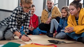 Eine Gruppe Kinder arbeitet zusammen auf dem Boden sitzend - inmitten eines männlichen Erziehers