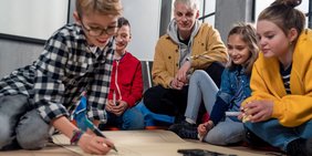 Eine Gruppe Kinder arbeitet zusammen auf dem Boden sitzend - inmitten eines männlichen Erziehers