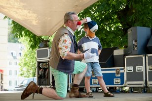 Willi mit einem Jungen beim clownesken Kindertheater auf der Bühne. Willi hält dem Jungen ein Basecap vor das Gesicht.