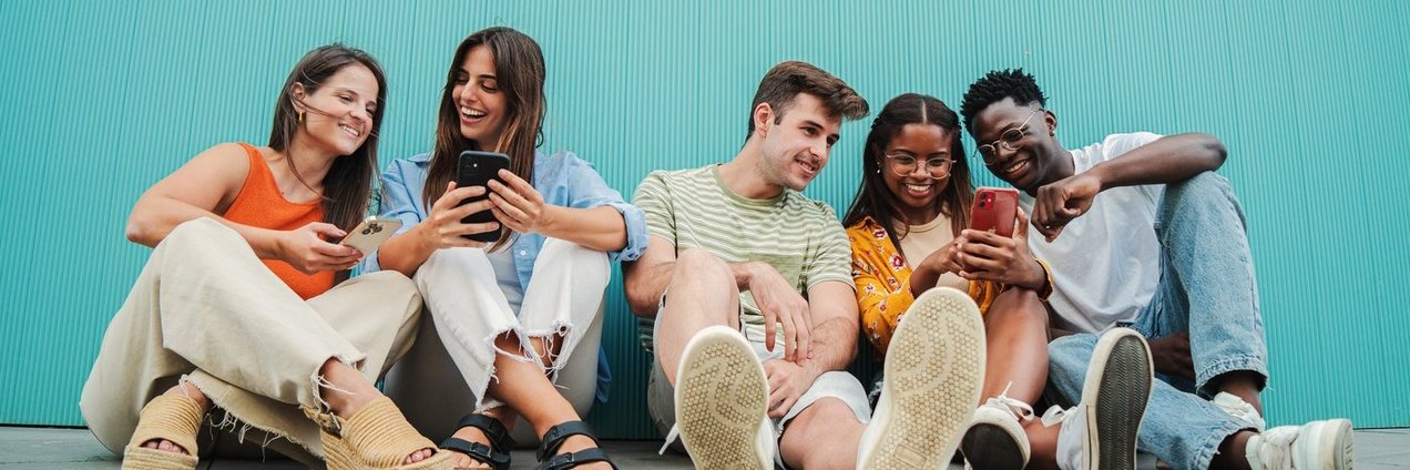5 Jugendliche mit 3 Smartphones sitzen an Wand gelehnt und schauen lächelnd auf ihre Smartphones