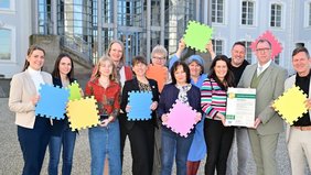 Regionalverbandsdirektorin Dr. Carolin Lehberger nimmt auf dem Schlossplatz die Auszeichnung als Gesundes Unternehmen für den Regionalverband entgegen. Neben Vertretern der AOK Rheinland-Pfalz/Saarland sind neben ihr auch Vertreterinnen und Vertreter auf dem Schlossplatz als Gruppe zusammengekommen. Alle halten sie bunte Puzzle-Stücke um das Zusammengreifen der verschiedenen Menschen für ein gelingendes BGM zu symbolisieren.