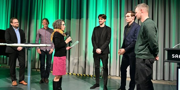 Drei Jurymitglieder laudieren die Gründungsmitglieder von fugeefilms gGmbH auf der Bühne des Festsaals des Saarbrücker Schlosses.
