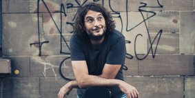 Manuel Sattler steht mit locker überkreuzten Armen vor einer mit Graffiti besprühten Mauer. Er trägt Jeans und ein dunkelblaues Shirt.