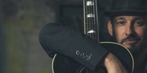 Pascal Geiser hinter seiner dunkelbraunen Gitarre vor verschwommenem Hintergrund.