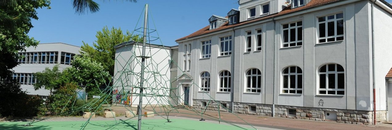 Ein helles Schulgebäude mit großen Fenstern davor ein Schulhof mit Kletternetz