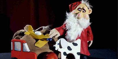 Die Figur eines Weihnachtsmanns. Neben ihm in einem kleinen Wagen ein Säckchen, vor ihm ein Apfel, eine Apfelsine, Walnüsse und die Holzfigur eines Hundes.