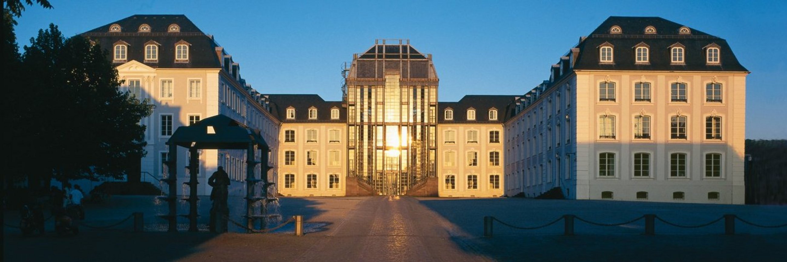 Blick auf das Saarbrücker Schloss mit Vorplatz. In der Glasfront spiegelt sich die untergehende Sonne