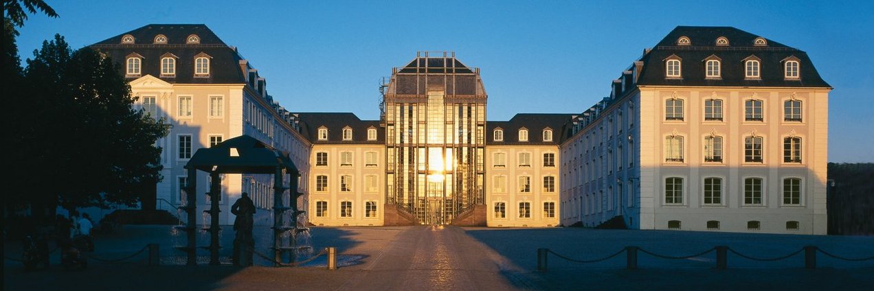 Blick auf das Saarbrücker Schloss mit Vorplatz. In der Glasfront spiegelt sich die untergehende Sonne