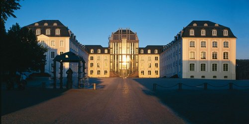 Blick auf das Saarbrücker Schloss mit Vorplatz. In der Glasfront spiegelt sich die untergehende Sonne