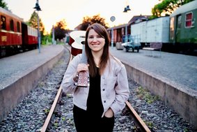 Eine junge Frau mit langem dunklem Haar steht bei fahlem Licht auf einem Bahngleis. Sie hält eine Gitarre, die auf ihrer Schulter ruht, und lächelt die Betrachtenden an.