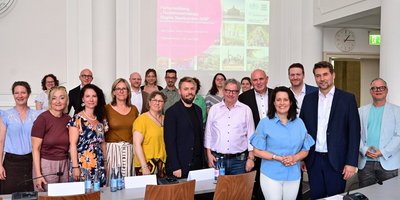 Eine Gruppe vor einer Projektion mit der Aufschrift "Fortschreibung Tourismusstrategie Region Saarbrücken 2030 lächelt im Sitzungssaal des Saarbrücker Rathauses in die Kamera