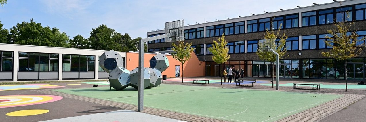 ein großer Schulhof mit bunten Bodenbemalungen, einem Sportfeld und einer Klettermöglichkeit vor einem hellen Schulgebäude mit großen Fenstern