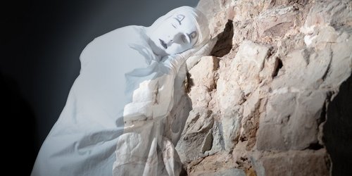 Das weiße, halb-durchsichtbare Schlossgespenst lehnt gegen eine Mauer im Schlosskeller und schläft.
