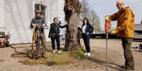 Die Baumscheibensanierung der Silberlinde begutachteten Regionalverbandsdirektorin Dr. Carolin Lehberger (2.v.r.) und Heike Schneider von der Aufbaugesellschaft Saarbrücker Schloss. Beide stehen um die Silberlinde herum. Ebenfalls auf dem Foto zwei ausführende Arbeiter, einmal mit Pressluftlanze und großem Bohrer