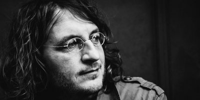 Schwarz-Weiß-Porträt von Manuel Sattler. Der Sänger hat schulterlanges, welliges Haar, trägt Brille und einen Dreitagebart.