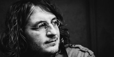 Manuel Sattler Schwarz-Weiß-Porträt von Manuel Sattler. Der Sänger hat schulterlanges, welliges Haar, trägt Brille und einen Dreitagebart.