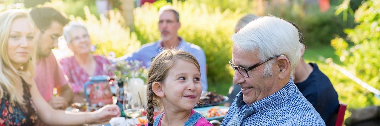 Eine Großfamilie feiert eine Gartenparty - im Vordergrund sitzt ein kleines Mädchen auf dem Schoß von ihrem Großvater