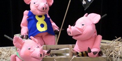 Drei rosafarbene Schweinchen-Figuren sitzen auf mit Stroh bedeckten Holzkisten.