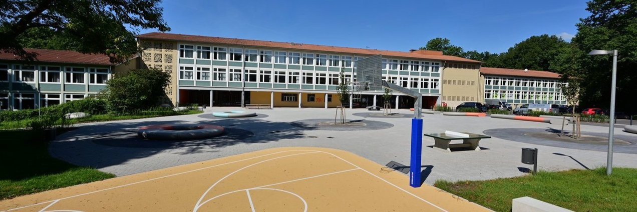 Schulhof mit Sportplatz mit Basketballkorb im Hintergrund ein großes Schulgebäude