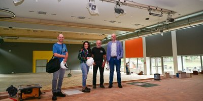 Christian Ackermann (Bausachbearbeiter), Regionalverbandsdirektorin Dr. Carolin Lehberger, Schulhausmeister Günter Kunkel und Bauamtsleiter Peter Koch stehen auf der Bühne in der Aula des Völklinger Marie-Luise-Kaschnitz-Gymnasiums. Im Hintergrund sind die Sanierungsarbeiten an der Aula zu sehen.