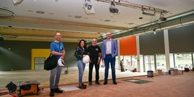 Christian Ackermann (Bausachbearbeiter), Regionalverbandsdirektorin Dr. Carolin Lehberger, Schulhausmeister Günter Kunkel und Bauamtsleiter Peter Koch stehen auf der Bühne in der Aula des Völklinger Marie-Luise-Kaschnitz-Gymnasiums. Im Hintergrund sind die Sanierungsarbeiten an der Aula zu sehen.
