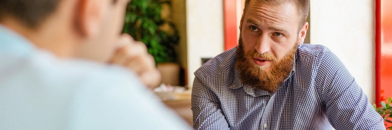 Blick über die Schuler eines Mannes, der am Tisch sitzt und mit einem jungen Mann ein Gespräch führt