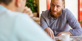 Blick über die Schuler eines Mannes, der am Tisch sitzt und mit einem jungen Mann ein Gespräch führt
