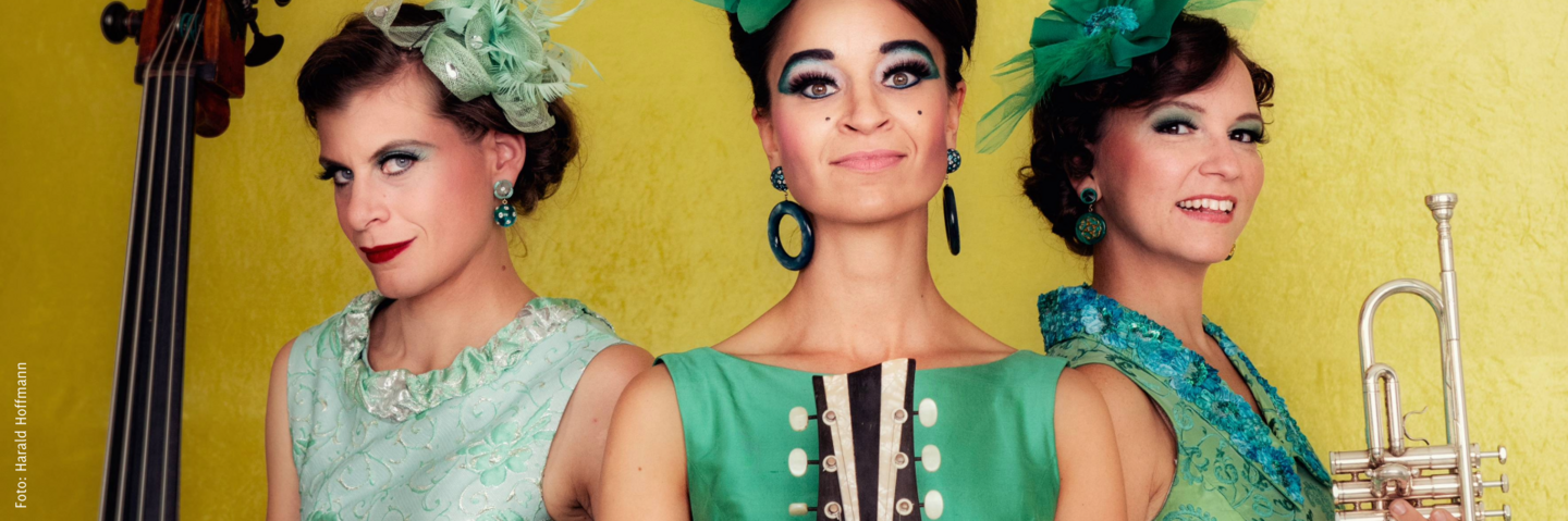 Die drei Musikerinnen der Zucchini Sistaz in grünen Kleidern und kleinen grünen Hüten vor einer gelben Wand stehend