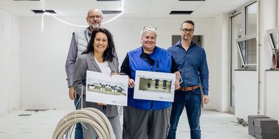 Vier Personen stehen im Rohbau der späteren Lehrküche und halten die Pläne der Küche in die Kamera. Auf dem Foto sind Regionalverbandsdirektorin Dr. Carolin Lehberger (v.l.), Bauamtsleiter Peter Koch (h.l.) und Mitarbeiter Marc Geber sowie Schulhausmeisterin Anja Arandt. 