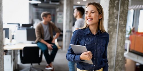 Junge fröhliche Mitarbeiterin mit Laptop in stylischen Büroräumen, im Hintergrund Kollegen im Gespräch