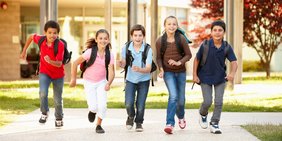Fünf Grundschulkinder verlassen mit ihren Ranzen die Schule und rennen der Kamera entgegen