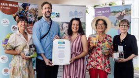 Fünf Menschen auf einer Bühne im Schlossgarten, die die Fairtrade-Urkunde und faire Produkte in die Kamera halten