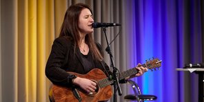 Die Sängerin Isabell Krohn tritt mit Gitarre auf der Bühne des Festsaals auf. Sie hat langes, dunkles glattes Haar und trägt einen schwarz-karierten Blazer.