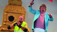 Auf einer Bühne: Ein Mann in Knieschonern und gelber Warnweste, neben ihm auf einem Hocker ein Mann mit geöffnetem, rotem Regenschirm.