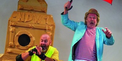 Auf einer Bühne: Ein Mann in Knieschonern und gelber Warnweste, neben ihm auf einem Hocker ein Mann mit geöffnetem, rotem Regenschirm.