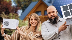 Eine Frau hält eine Miniaturnachbildung einer Wärmepumpe in die Luft. Neben ihr steht ein Mann, der eine Miniaturnachbildung eines Hauses mit einer Solaranlage auf dem Dach präsentiert. Beide abgelichtet vor einem Einfamilienhaus im Grünen.