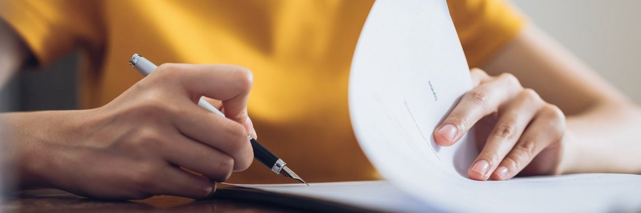 Woman signing document and hand holding pen putting signature at Eine Frau unterschreibt ein Dokument; ihre Hand hält einen Stift und setzt die Unterschrift auf das Dokument.