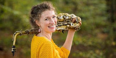 Nicole Johänntgen, eine circa vierzigjährige Musikerin, lächelt den Betrachter von der Seite an. Sie hält ihr goldfarbenes Saxophon, das auf ihrer linken Schulter ruht.