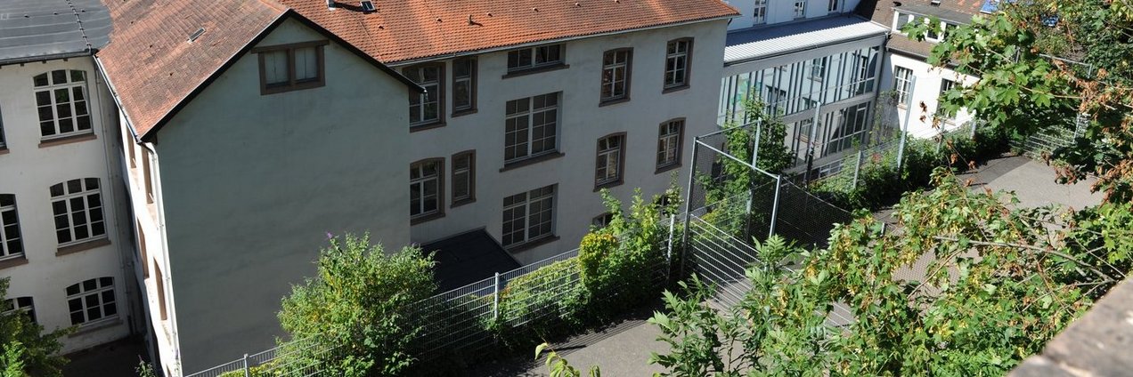 Blick auf den oberen Hof des Gymnasiums am Schloss in Saarbrücken mit historischem Schulgebäude, Tischtennisplatte, Bäumen und sonnigem Himmel.