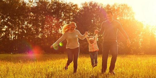 Familie läuft Hände haltend über ein Feld. Im Hintergrund geht die Sonne unter.